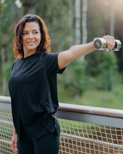 Active energized sportswoman stretches arms with dumbbell, poses on bridge over nature background with sunrise in morning, dressed in active wear, looks into distance, works on training biceps